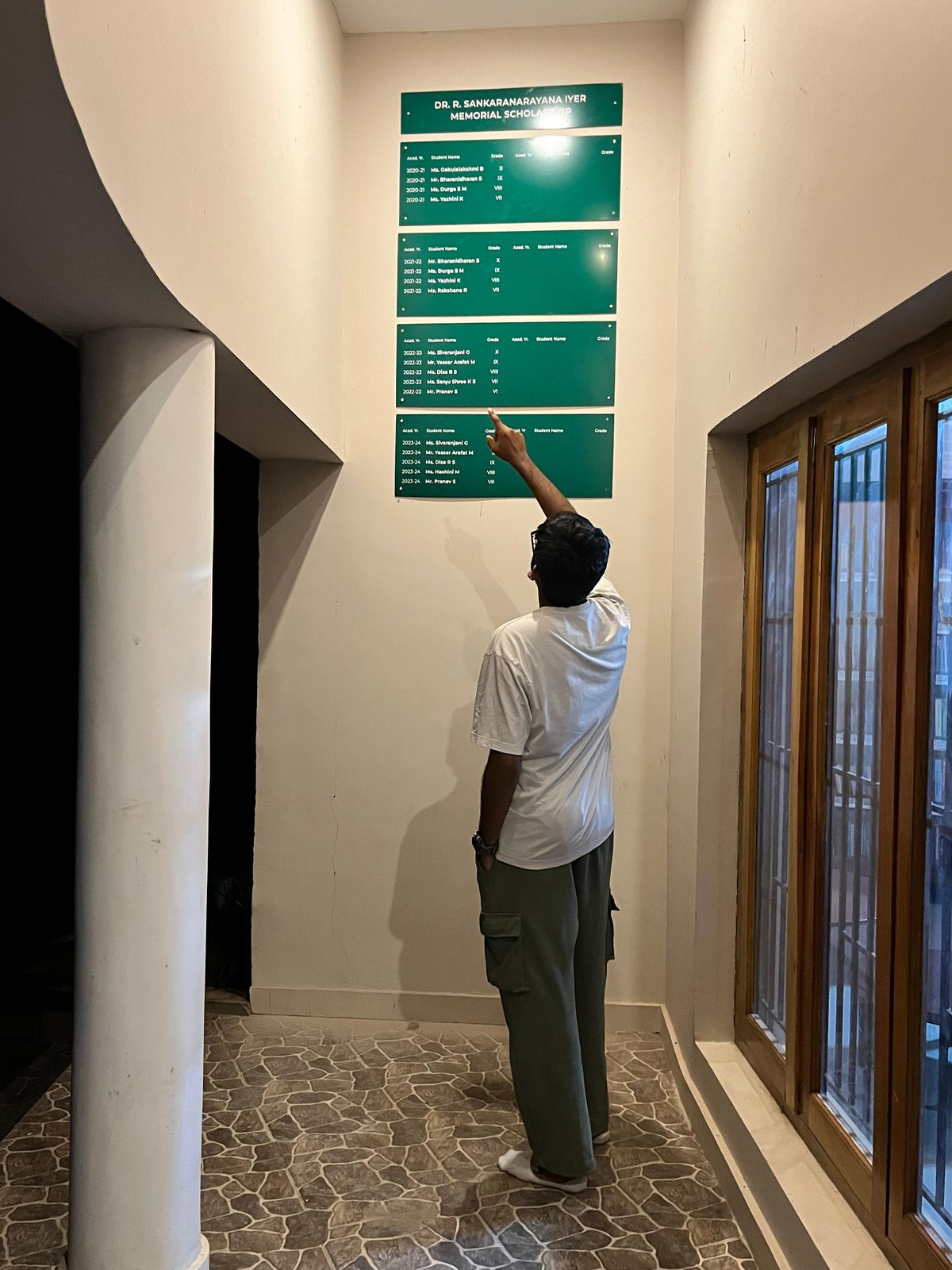 The finished green Sankaranarayana Iyer Memorial Scholarship board installed on the wall, with Akash pointing up at a name